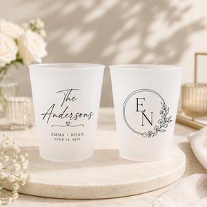 May include: Two frosted plastic cups featuring black printed text. One cup displays The Andersons, Emma + Noah, and June 18, 2028. The other cup features the initials E N inside a circular frame decorated with a delicate floral vine illustration.
