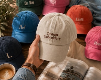 May include: A collection of baseball caps in various colors including beige, blue, green, pink, and orange. The beige cap in the foreground features the text Custom Embroidery in brown thread. Other caps display embroidered words like Cat Mom, Yes, No, Esme, and Blessed.
