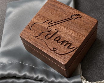 May include: A square dark brown wooden box with a hinged lid featuring an engraved outline of an electric guitar and the word Jam in cursive script. The box is positioned on a smooth, light gray satin drawstring pouch with a matching ribbon tie.