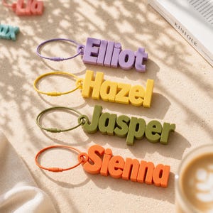May include: Four custom keychains featuring 3D block lettering in lavender, yellow, olive green, and orange. Each keychain includes a matching circular metal wire ring with a screw-top closure. The names Elliot, Hazel, Jasper, and Sienna are displayed.