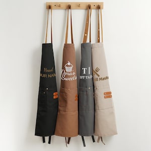May include: Four aprons hanging on a wooden rack. The aprons are black, brown, grey, and beige. Each apron has a pocket and leather straps. Text on the aprons includes "Head OUR NAM", "Dora's Sweets", "T | ST TAD", and "Grill Master".