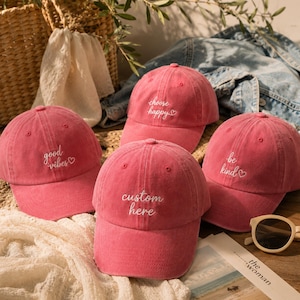 Peut inclure: Quatre casquettes de baseball roses avec du texte brodé en blanc. Les casquettes portent les inscriptions "good vibes", "choose happy", "custom here" et "be kind", chacune accompagnée d'un petit cœur. Elles ont un aspect délavé.