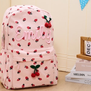 Op de afbeelding: Roze corduroy rugzak met een aardbeienpatroon en de naam "Emily" in roze letters. De rugzak heeft een voorvak en is versierd met kersen en bladeren. Een stijlvol en functioneel accessoire voor school of uitstapjes.