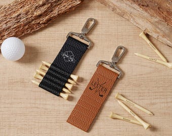 May include: A golf-themed product arrangement featuring a white golf ball, two leather tee holders with metal clasps, and personalized wooden golf tees. One holder is black, the other is brown, both with golf-related graphics.