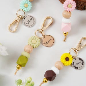 Op de afbeelding: Sleutelhangers met decoratieve kralen en bloemaccenten. Elke sleutelhanger heeft een goudkleurige sluiting, houten kralen en een bedel met een naam. De sleutelhangers hebben verschillende kleuren, waaronder groen, roze, geel en bruin.