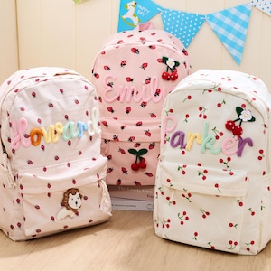 Op de afbeelding: Drie corduroy rugzakken met een aardbeien- en kersenpatroon. Eén rugzak heeft de naam "Howard" in regenboogletters, een andere "Emil" en de derde "Parker". Elke rugzak heeft een voorvak.