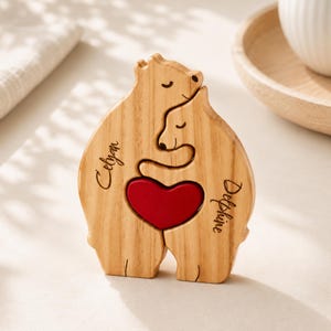 Op de afbeelding: Een lichtbruin houten berenbeeldje met twee beren die elkaar omhelzen, met een rood hart in het midden. De naam Celyan is gegraveerd aan de linkerkant en de naam Delphine aan de rechterkant van dit houten berenbeeldhouwwerk.