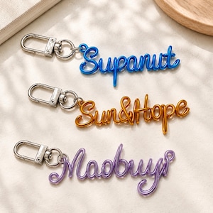 May include: Three metal keychains with silver-toned lobster clasps. Each features a different word formed from continuous wire: the blue one reads Supanut, the gold one reads Sun&Hope, and the purple one reads Maobuy in a cursive script.