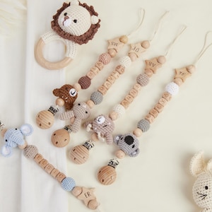 Op de afbeelding: Een verzameling babyaccessoires, waaronder gehaakte dierentanden en fopspeenclips. De fopspeenclips hebben houten kralen met gepersonaliseerde namen en gehaakte dierenfiguren in verschillende kleuren. Een gehaakte leeuwentand en een konijn zijn ook zichtbaar.