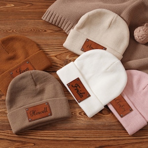 Puede incluir: Cinco gorros de punto en varios colores: marrón, beige, blanco y rosa. Cada gorro tiene un parche rectangular de cuero marrón con un nombre grabado. Los gorros están colocados sobre una superficie de madera.