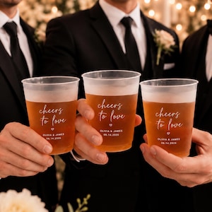 May include: Three clear plastic cups filled with amber liquid, each printed with the white text cheers to love, a small heart, and the names Olivia & James with the date 14.06.2026, held by individuals wearing black suits and black ties.