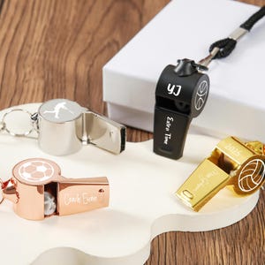 May include: A selection of sports whistles in silver, black, and gold. The whistles are designed for soccer, basketball, and volleyball. Some whistles have text that reads "Extra Time", "Coach Evan", and "2025".