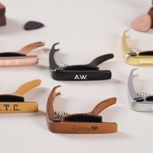 Op de afbeelding: Een verzameling gitaarcapo's in diverse kleuren en materialen, waaronder roségoud, zwart, goud, zilver en hout. Elke capo heeft een uniek ontwerp en gepersonaliseerde tekst, zoals initialen of een naam. De capo's worden gepresenteerd op een witte ondergrond.