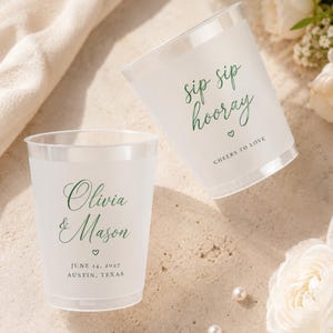May include: Two frosted plastic cups featuring green script text. One cup displays "sip sip hooray" with a heart and "CHEERS TO LOVE". The other cup shows "Olivia & Mason" with a heart, "JUNE 14, 2027", and "AUSTIN, TEXAS" in black text.