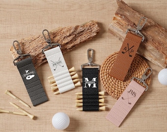 May include: Leather golf keychains in black, white, brown, and pink, each with a silver clasp and attached golf tees. The keychains are personalized with initials or names, and feature golf club graphics. Two golf balls are also visible.