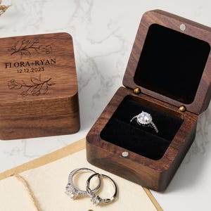 May include: A wooden ring box with a hinged lid. The box is engraved with the text "FLORA + RYAN 12.12.2023" and a floral design. The box is open and a ring is inside.