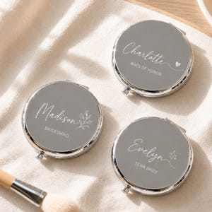 Op de afbeelding: Drie ronde, zilverkleurige compacte spiegels met grijze deksels. Elk deksel is voorzien van witte aangepaste tekst en afbeeldingen: Charlotte met een hartje, Madison met een blaadje en Evelyn met een ster. De tekst bevat namen en titels.