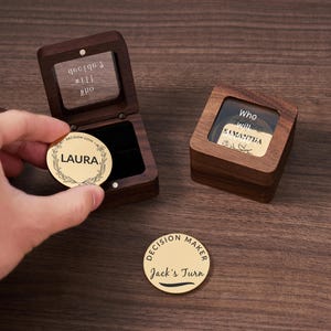 May include: Two wooden boxes with a gold coin inside. The coin in the left box has the word "LAURA" engraved on it. The coin in the right box has the words "Who will Samantha" engraved on it. The box on the right also has a glass lid. The coin on the table says "Decision Maker Jack's Turn".