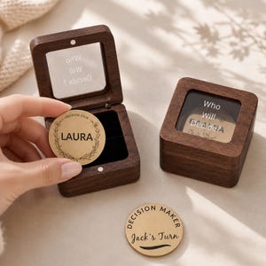 May include: Two dark brown wooden boxes with glass lids featuring the text Who Will Decide? and two circular metal decision coins. One coin is held showing the name LAURA inside a floral wreath, and the other coin reads DECISION MAKER Jack's Turn.