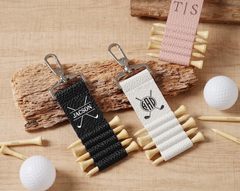 May include: Golf tee holders in black, white, and pink leather with silver keychains. The black holder has "JACSON" printed on it, and the white holder has a golf club logo. A golf ball and tees are also present.