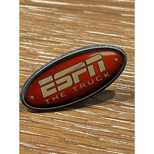 May include: An oval-shaped pin with a red background and the ESPN logo in gold. The text "THE TRUCK" is below the logo. The pin has a silver border and is set against a wooden surface.