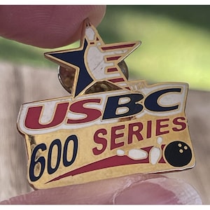 May include: A gold-coloured pin featuring the text "USBC 600 SERIES" with a bowling ball graphic. The pin also includes a star design with red, white, and blue accents, and the USBC logo.