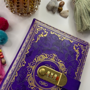Peut inclure: Journal en cuir violet avec un motif en relief doré et une serrure à combinaison. Le journal a une fermeture à sangle et est décoré d'un motif floral doré.
