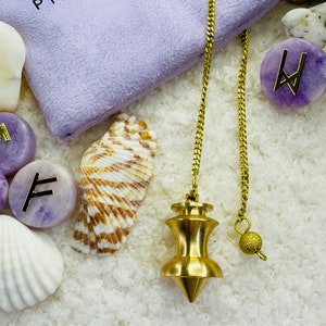 May include: A gold-colored pendulum with a pointed tip hangs from a gold chain. The pendulum is on a white fluffy surface with purple and white stones, a seashell, and a purple velvet bag with the text "PI.SPIRITUEL" on it.