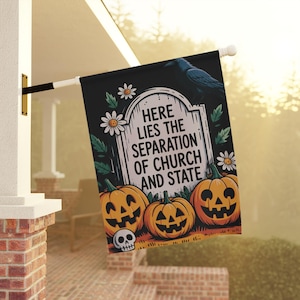 May include: A black rectangular garden flag featuring a central tombstone with the text 'HERE LIES THE SEPARATION OF CHURCH AND STATE'. The design includes three orange jack-o'-lanterns, a small skull, white daisies, and a black raven at the top.