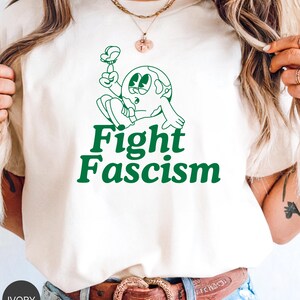 Puede incluir: Camiseta de color marfil con un personaje de dibujos animados verde sosteniendo una flor y las palabras "Fight Fascism" en verde. La camiseta tiene cuello redondo y mangas cortas. La modelo lleva un cinturón marrón con una hebilla decorativa.