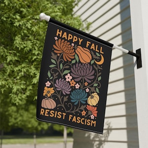 Può includere: Bandiera nera da esterno con le parole "HAPPY FALL" e "RESIST FASCISM" in arancione. La bandiera presenta un disegno floreale con zucche, fiori e una luna crescente nei toni dell'arancione, viola e blu.