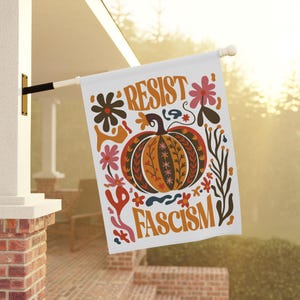 Può includere: Bandiera bianca da esterno con le parole "RESIST FASCISM" in lettere arancioni. La bandiera presenta un design decorativo di zucca e floreale nei toni dell'arancione, marrone e rosso. La bandiera è appesa a un palo bianco.