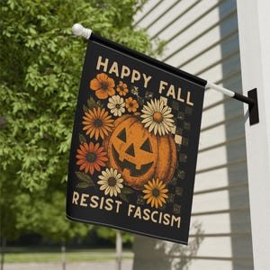 May include: Black outdoor flag with the text "HAPPY FALL RESIST FASCISM". The flag features a smiling jack-o'-lantern surrounded by orange and white flowers. The background has a checkered pattern.