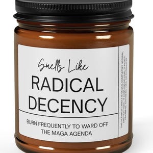Op de afbeelding: Een bruine glazen potkaars met een zwart deksel. Het etiket luidt "Smells Like RADICAL DECENCY" en "BURN FREQUENTLY TO WARD OFF THE MAGA AGENDA". De kaars is veganistisch, natuurlijk en niet giftig.