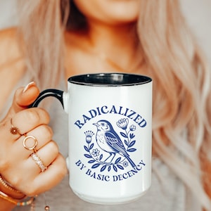 May include: White ceramic mug with a black handle and rim. The mug features a blue illustration of a bird surrounded by flowers and the text "RADICALIZED BY BASIC DECENCY" in a blue, stylized font.