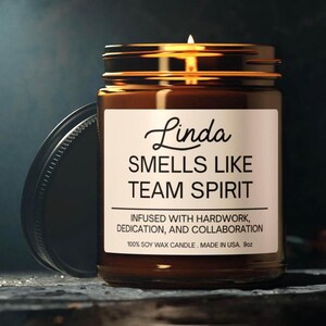 以下が含まれることがあります： 255gのアンバーガラス瓶入りキャンドル。白いラベルには「Linda SMELLS LIKE TEAM SPIRIT INFUSED WITH HARDWORK, DEDICATION, AND COLLABORATION 100% SOY WAX CANDLE. MADE IN USA.」と記載されています。火のついた芯と黒い蓋が付いています。