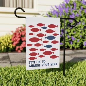 Può includere: Una bandiera da giardino bianca con un motivo di pesci rossi e blu. La bandiera presenta il testo "IT'S OK TO CHANGE YOUR MIND" in blu navy. La bandiera è esposta su un supporto in metallo nero in un giardino.