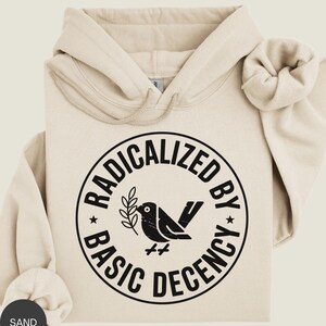 May include: Sand-colored hoodie with a drawstring hood and a large graphic on the front. The graphic is a black circle with the words "RADICALIZED BY BASIC DECENCY" surrounding a black bird holding a sprig. The hoodie is folded.