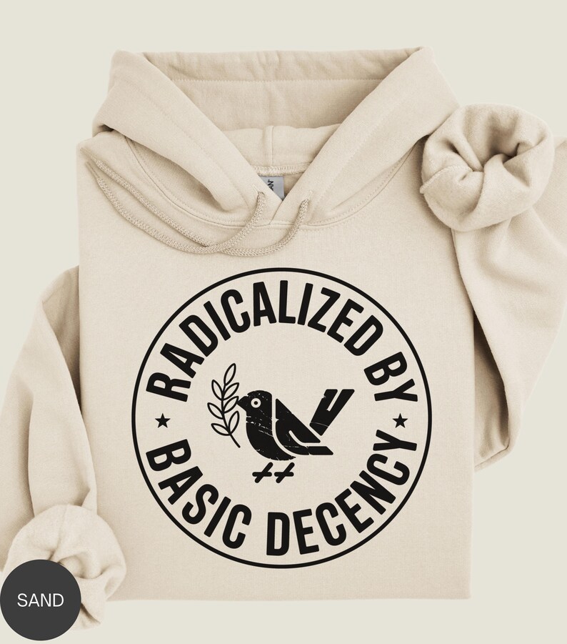 May include: Sand-colored hoodie with a drawstring hood and a large graphic on the front. The graphic is a black circle with the words "RADICALIZED BY BASIC DECENCY" surrounding a black bird holding a sprig. The hoodie is folded.