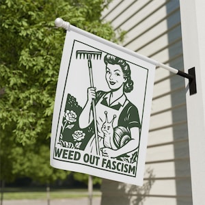 May include: A white flag with a green illustration of a woman holding a rake and a snail, with the text "WEED OUT FASCISM". The flag is mounted on a white pole and is displayed outdoors.