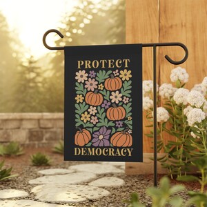 Peut inclure: Drapeau de jardin noir avec les mots "PROTECT DEMOCRACY" en lettres dorées. Le drapeau présente un motif floral avec des citrouilles orange, des feuilles vertes et des fleurs colorées. Le drapeau est affiché sur un piquet de jardin en métal noir.