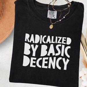 Puede incluir: Camiseta negra con la frase blanca «RADICALIZED BY BASIC DECENCY». Un collar de cuentas con un colgante descansa sobre la camiseta doblada, presentada sobre un fondo neutro.