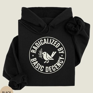 May include: Black hoodie with a white circular graphic that reads "RADICALIZED BY BASIC DECENCY" around a bird holding a sprig. The hoodie has a drawstring hood and long sleeves. The word "BLACK" is in a tan circle in the lower left corner.