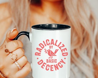 May include: White ceramic mug with a black handle and rim, featuring the text "RADICALIZED BY BASIC DECENCY" in red. A red bird graphic is in the center. The mug is held by a person with blonde hair and multiple rings.