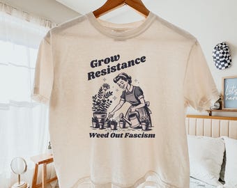Op de afbeelding: Crèmekleurig T-shirt met een zwart-witte afbeelding van een vrouw die planten water geeft, met de tekst "Grow Resistance" en "Weed Out Fascism". Het T-shirt hangt aan een houten hanger.