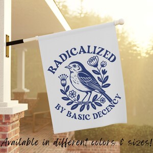 Pode incluir: Bandeira branca com um desenho azul marinho com um pássaro, flores e o texto "RADICALIZED BY BASIC DECENCY". A bandeira está pendurada em um mastro branco, montado em uma estrutura de tijolo.
