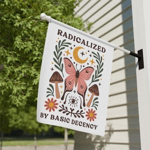 Op de afbeelding: Witte vlag met de tekst "RADICALIZED BY BASIC DECENCY" en een vlinder, paddenstoelen, maan, sterren en bloemenontwerp. De vlinder is roze met zwarte details. De vlag hangt buiten.