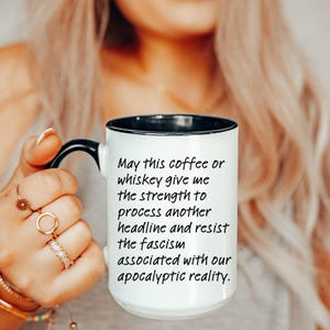 May include: A white ceramic mug with a black interior and handle, featuring the black printed text: May this coffee or whiskey give me the strength to process another headline and resist the fascism associated with our apocalyptic reality.
