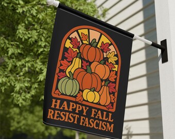 Puede incluir: Bandera negra con un diseño otoñal con calabazas y hojas de otoño en naranja, amarillo y verde. El texto "HAPPY FALL RESIST FASCISM" se muestra en naranja. La bandera está montada en un mástil blanco y negro.