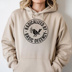 May include: Beige hoodie with a black circular graphic that reads "RADICALIZED BY BASIC DECENCY" around a bird holding a sprig. The hoodie has a drawstring hood and a front pocket.